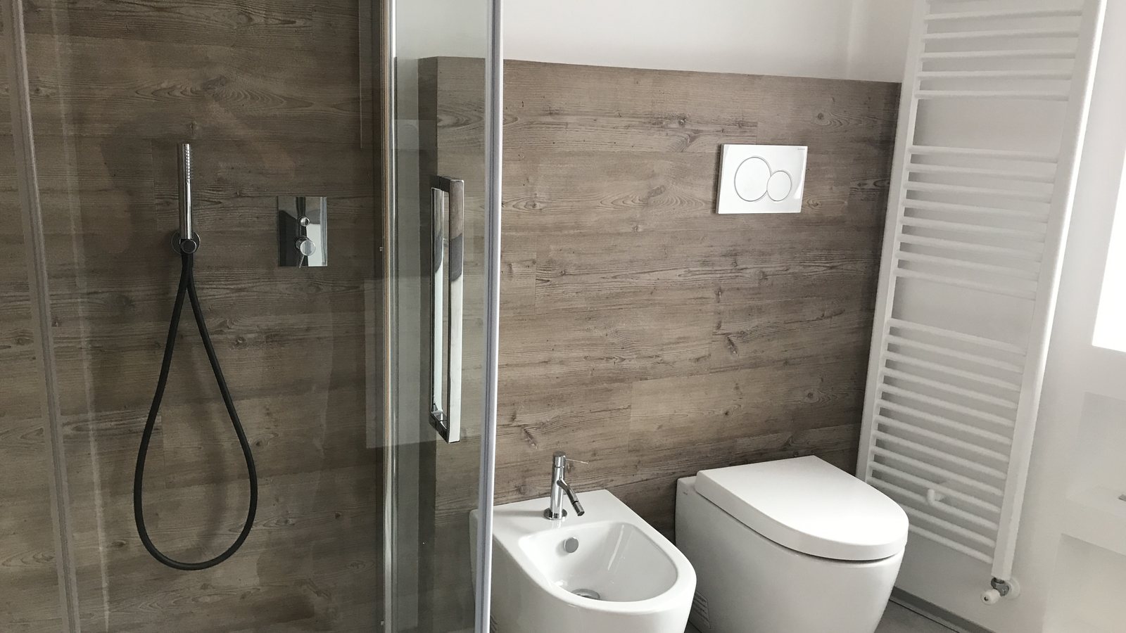 Bagno a Roncone dopo l'intervento con doccia in vetro, rivestimenti effetto legno e sanitari contemporanei