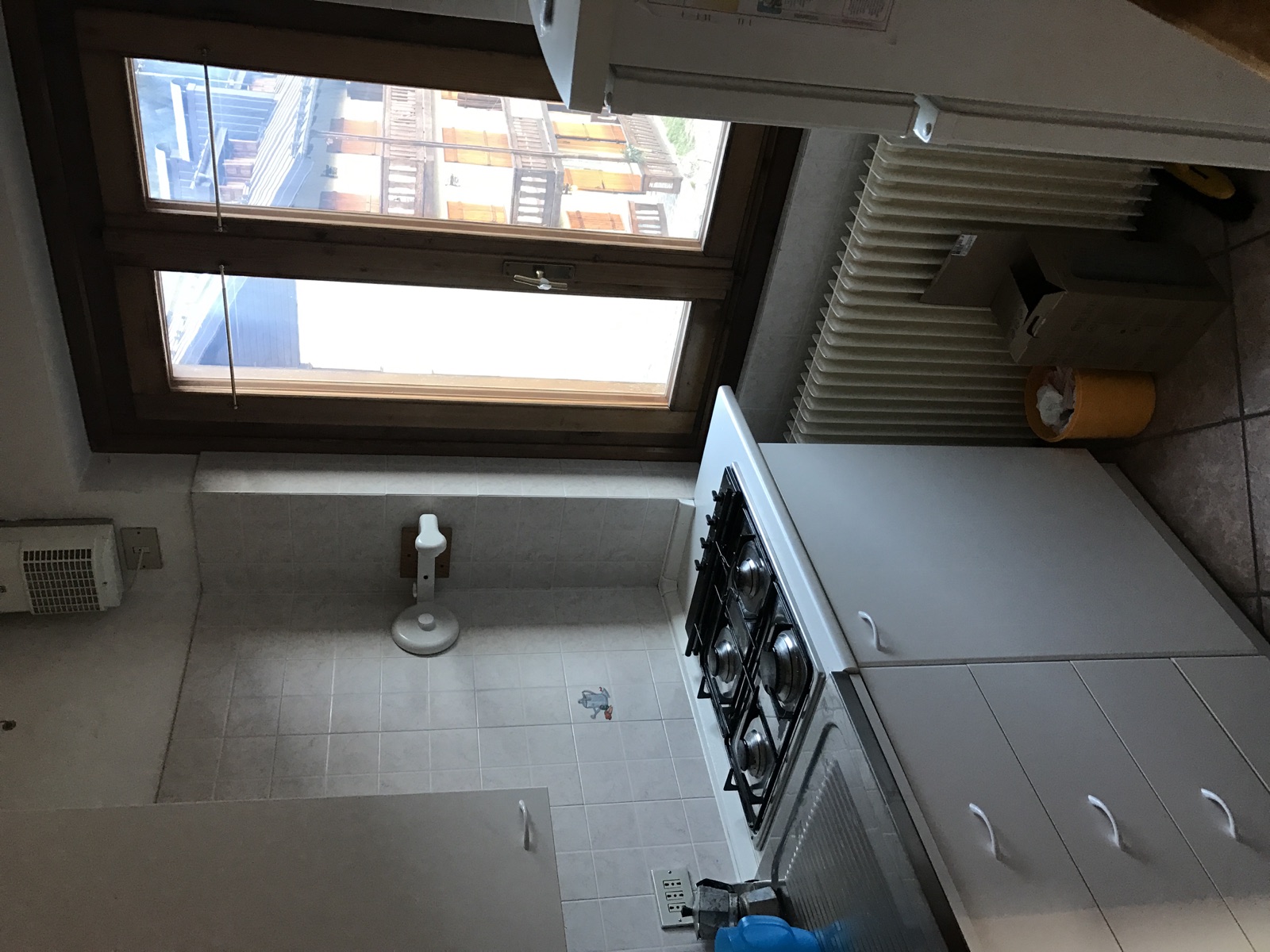 Cucina a Pinzolo prima dell'intervento con mobili bianchi, finiture datate e radiatore sotto la finestra